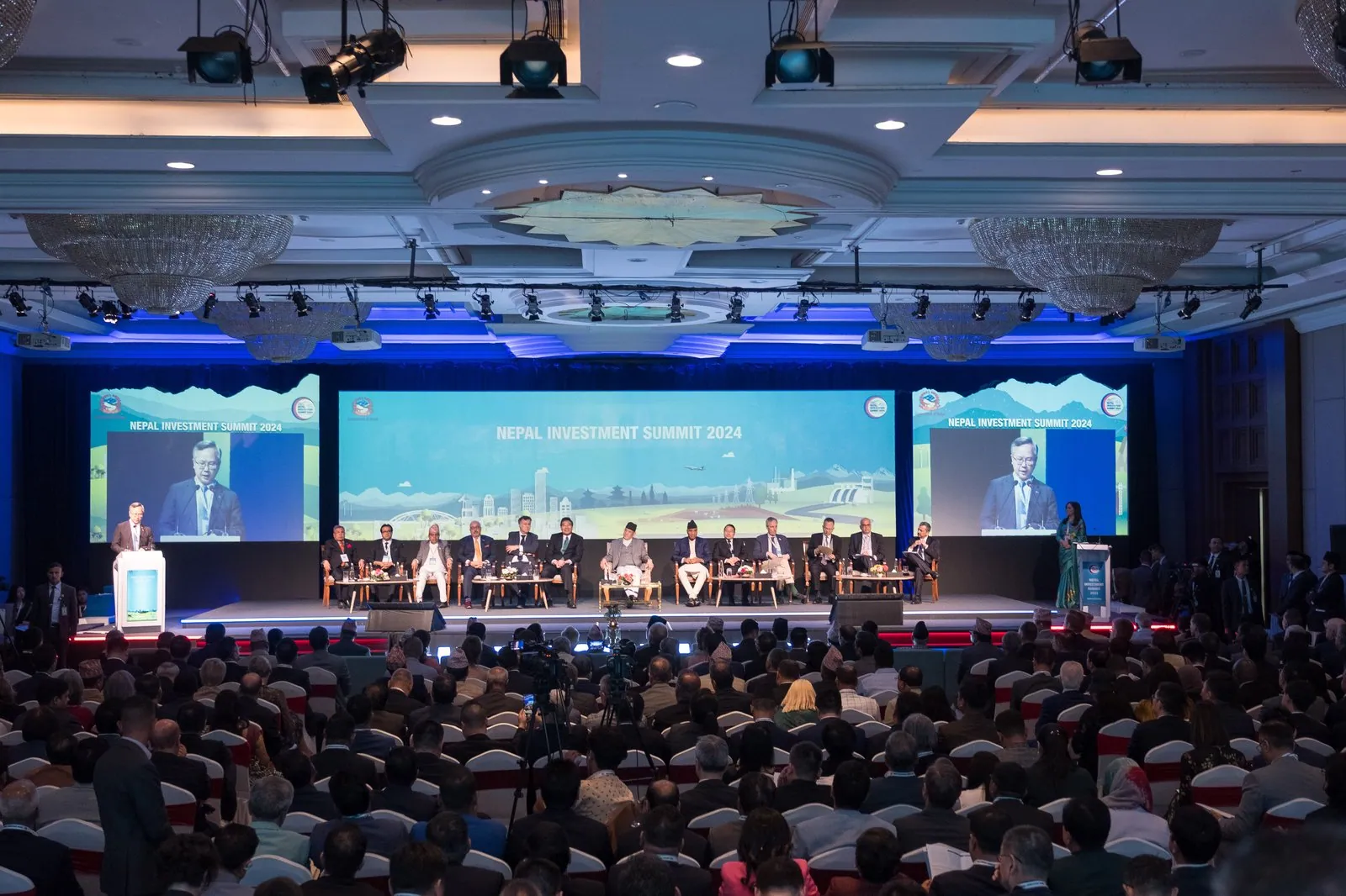A wide view of the Nepal Investment Summit 2024 hall: three screens above a stage with a row of seated guests, and a full audience in the foreground.