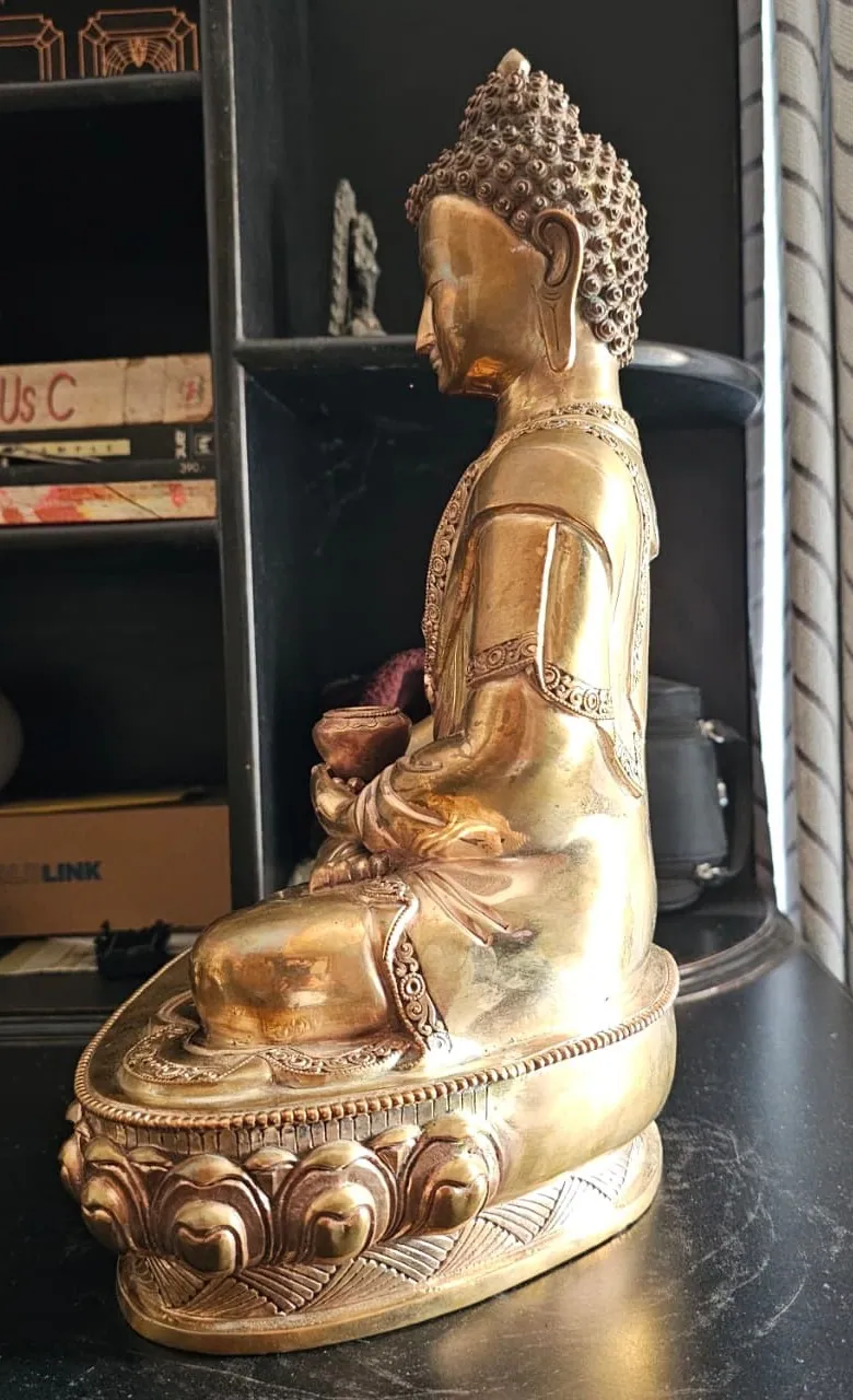 Profile view of the brass Buddha from the left, showing the alms bowl in the hands and the tiered lotus base.