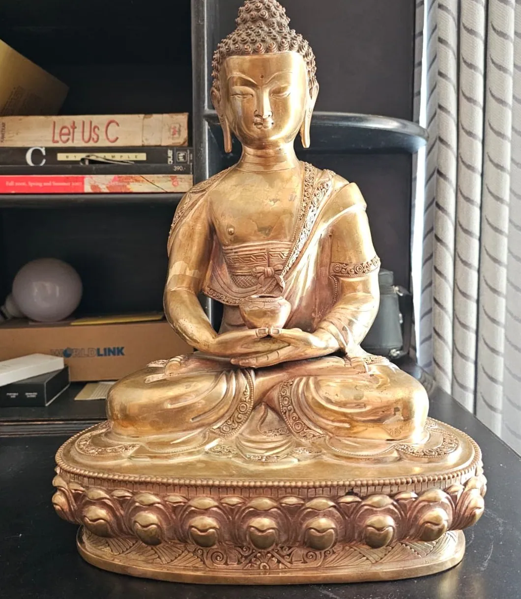 Front view of the polished brass Buddha seated on a lotus base, hands in meditation mudra holding an alms bowl.