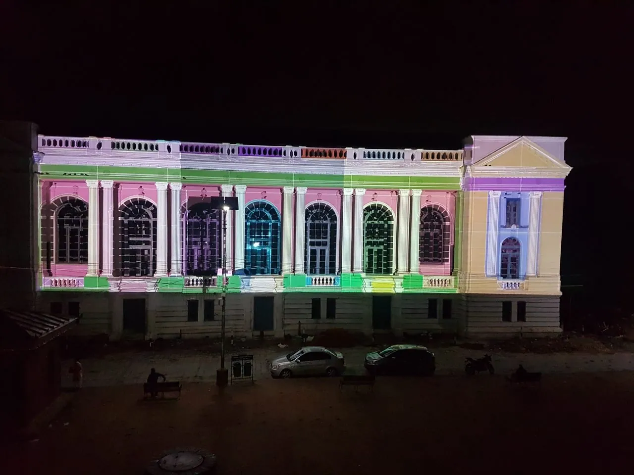 Night photograph of the facade from above, with pink, green, purple and cyan colour blocks and thin grid lines projected onto its walls, windows, frieze and plinth.