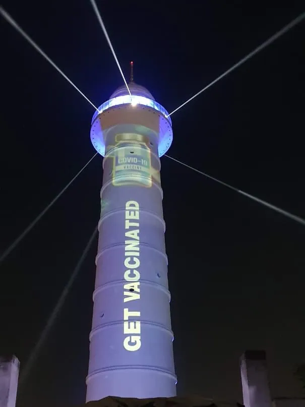 The tower at night with white laser beams and the words Get Vaccinated in large letters down the shaft, above a COVID-19 vaccine bottle.