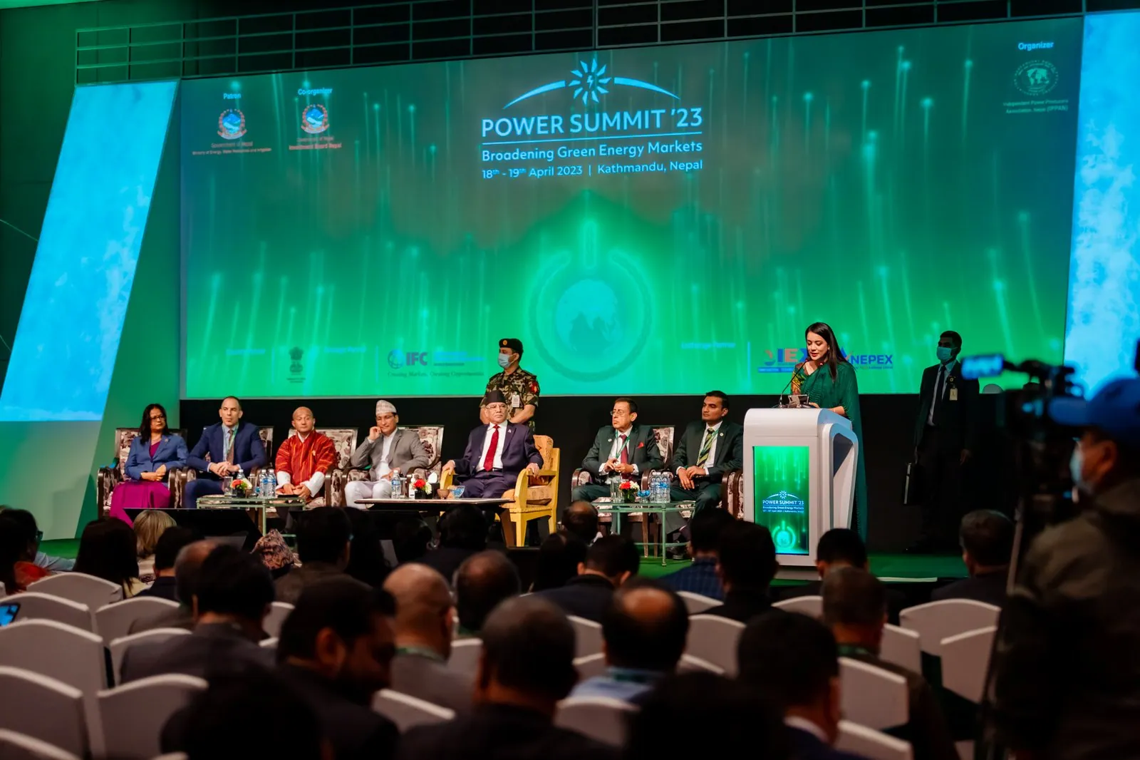 Power Summit '23 main stage: a green LED screen with the summit title and dates, seven guests seated on stage and a speaker at a white lectern.
