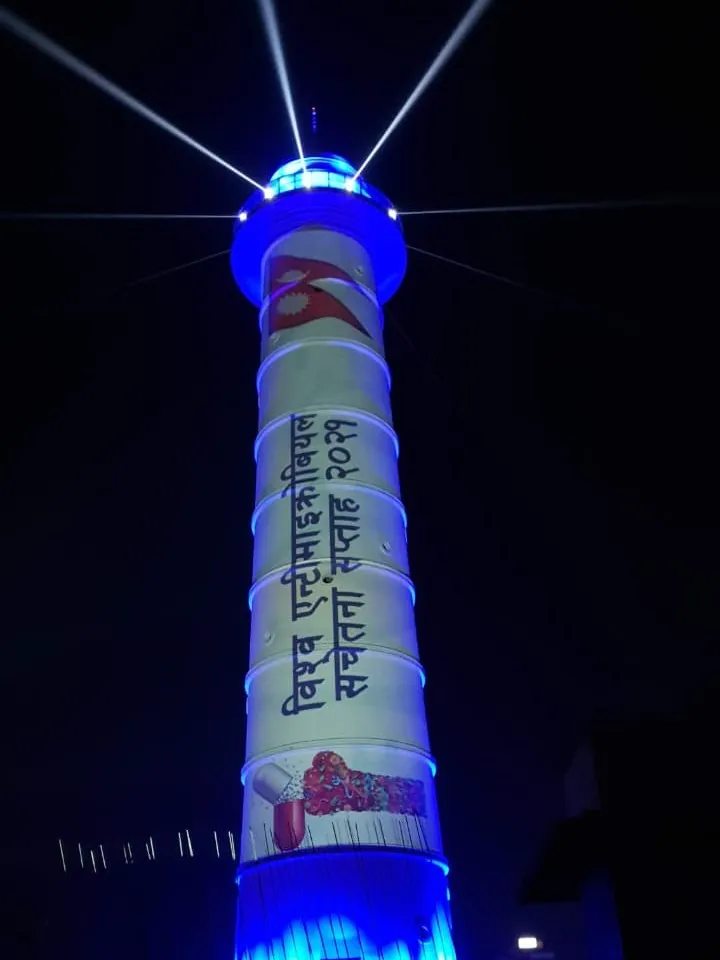 The tower in blue light with the Nepali title of World Antimicrobial Awareness Week 2021, the Nepali flag and a capsule graphic projected down the shaft.