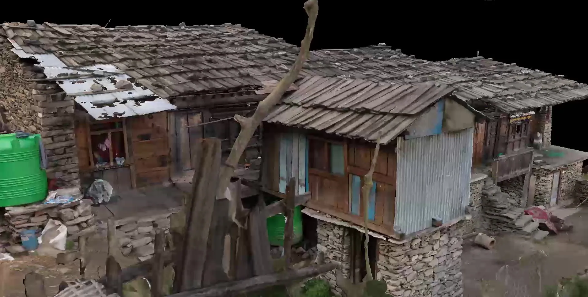 View of a timber house corner in the Gatlang model with a corrugated-metal wall, a green water tank and a leaning tree trunk.