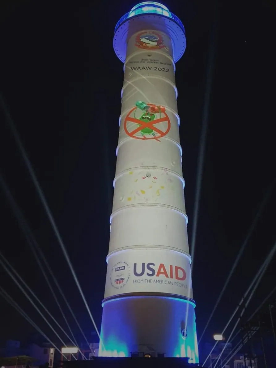 The tower in white with laser beams fanning out, showing the WAAW 2022 title, a crossed-out germ graphic and the USAID logo at the base.
