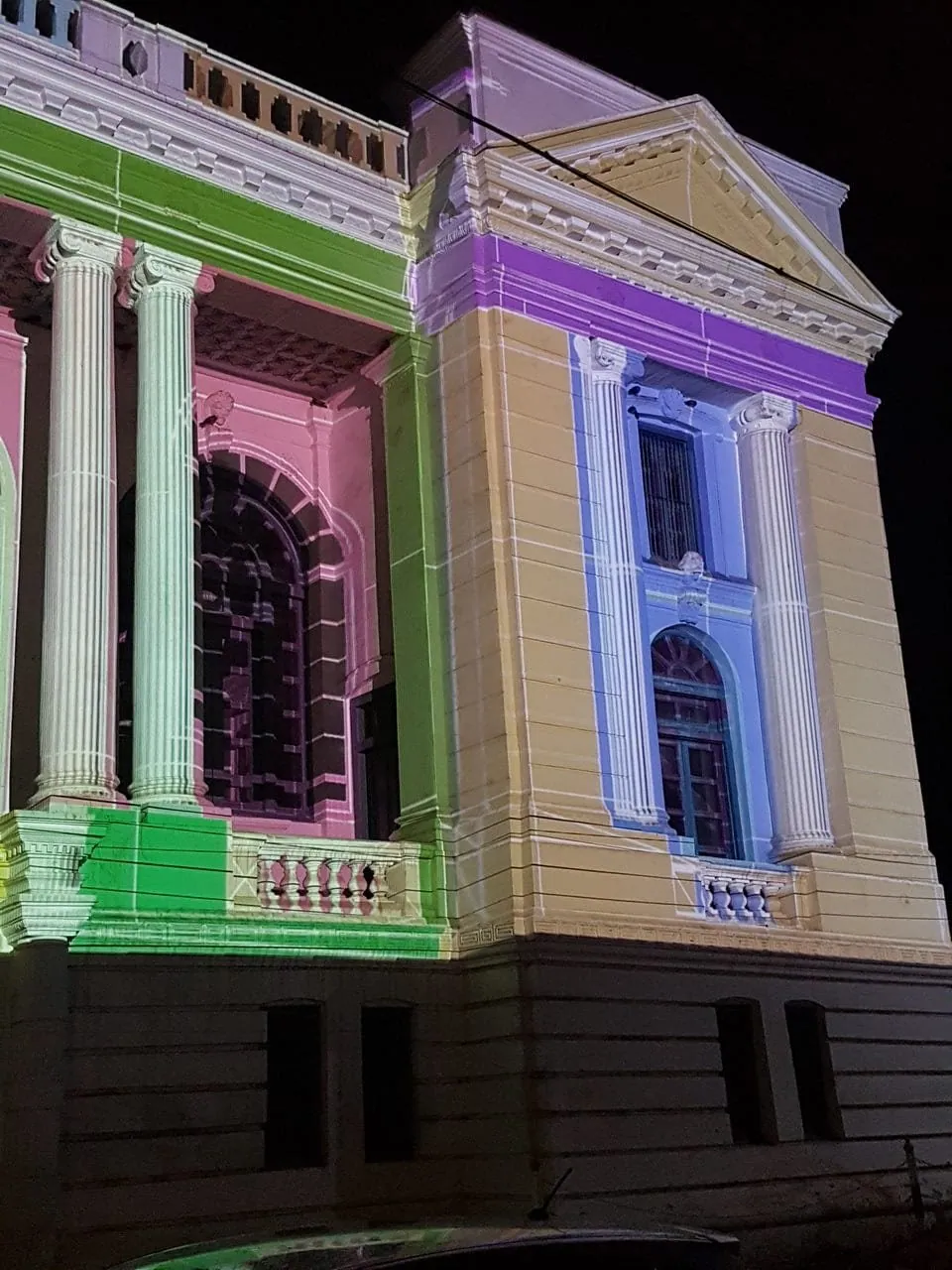 Close night photograph of a pavilion with a purple frieze band, blue-lavender window surround, green plinth block and pink wall, projected onto the stonework.
