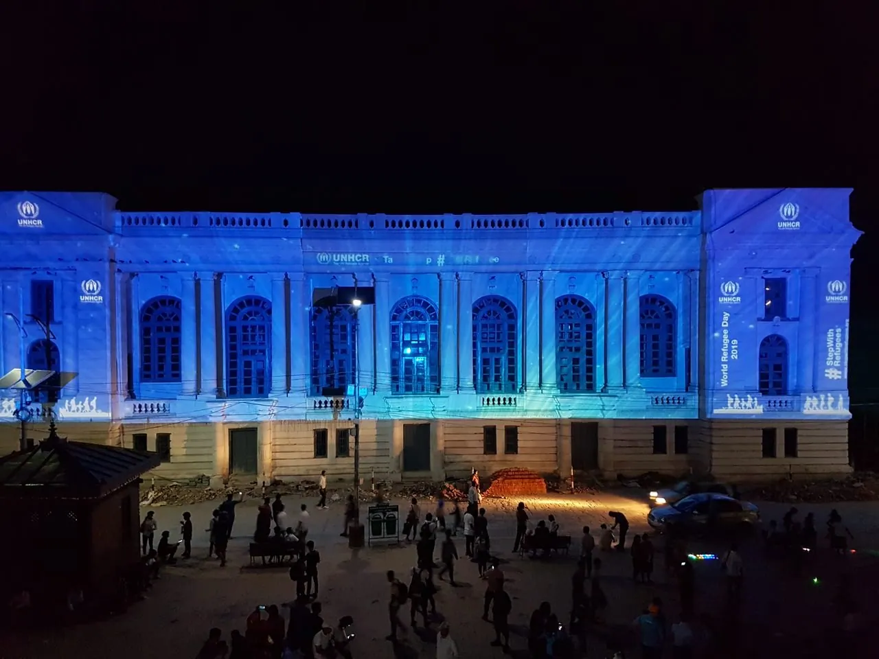 Night photograph of the facade with “World Refugee Day 2019” and “#StepWithRefugees” panels on the right pavilion, and the frieze text partly revealed.