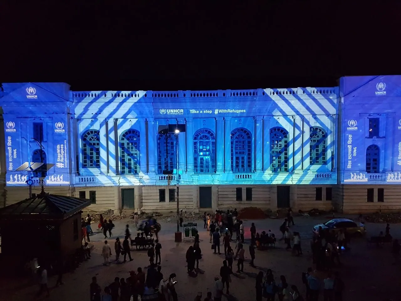 Night photograph of the facade in UNHCR blue with white diagonal bands crossing the walls and UNHCR logos on the pavilions, seen across a crowded square.